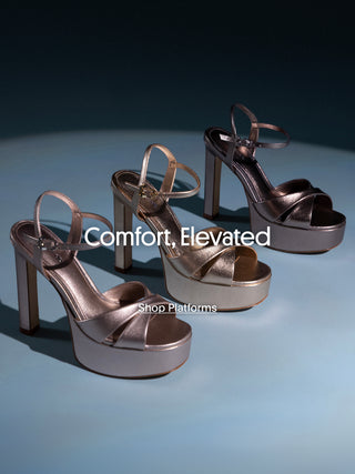Studio image of metallic platform high-heel sandals with ankle straps in silver and champagne tones, arranged on a blue surface against a dark background. White text over the image reads “Comfort, Elevated” with a smaller caption “Shop Platforms".