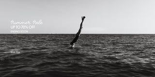 Person diving headfirst into the ocean while wearing black platform heels, with a monochrome seascape in the background.