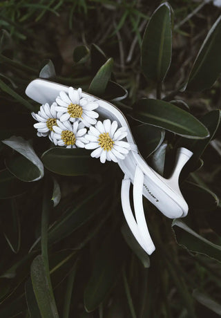 White leather slingback kitten heel adorned with white daisy embellishments, resting among lush green foliage in a moody, nature-inspired setting.