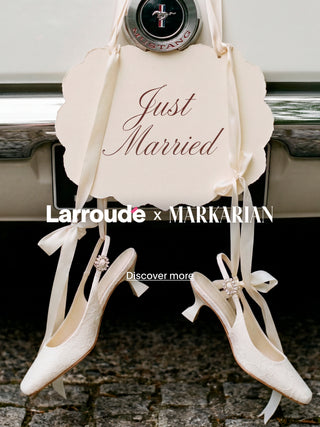 Pair of ivory bridal heels with satin ribbon ties and crystal embellishments hanging from a vintage car bumper with a 'Just Married' sign, featuring the Larroudé x Markarian collaboration logo and a 'Discover more' call-to-action.