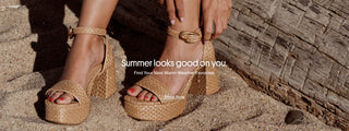 Larroudé raffia platform sandals with ankle straps and block heels, worn on a sandy beach beside driftwood.